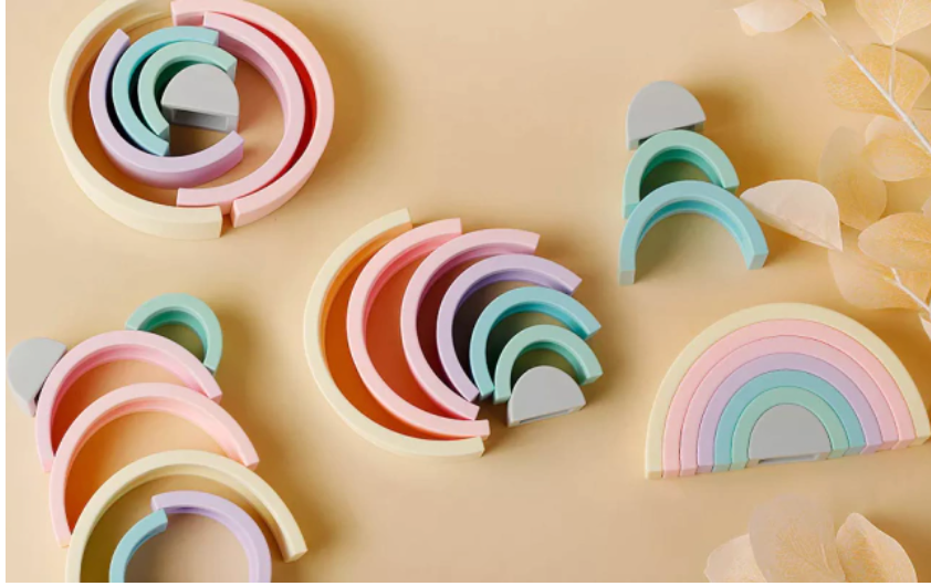 Arco-íris feito de tiras de silicone nas cores cinza, verde água, azul, roxo, lilás, laranja claro e amarelo claro. Peças desmontáveis para brincar e empilhar e encaixar. Peças colocadas de outras formas para ilustrar outras modos de brincar.
