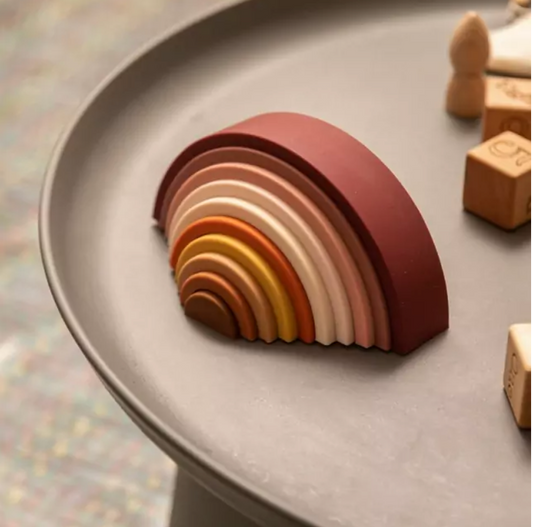 Arco-íris feito de tiras de silicone nas cores marrom, rosa, amarelo mostarda e laranja sobre uma mesa cinza ao lado de blocos de madeira. Peças desmontáveis para brincar e empilhar e encaixar.