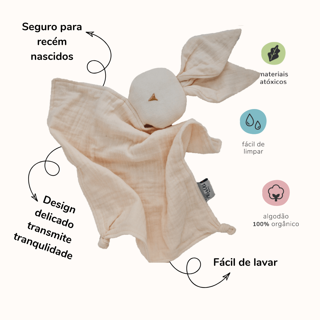 Naninha de algodão em formato de coelho na cor creme com informações sobre produtos. Do lado esquerdo da naninha as seguintes informações: Seguro para recém nascidos, design delicado, transmite tranquilidade. Do lado direito da naninha: materiais átoxicos, fácil de limpar, algodão 100% orgânico, fácil de lavar.