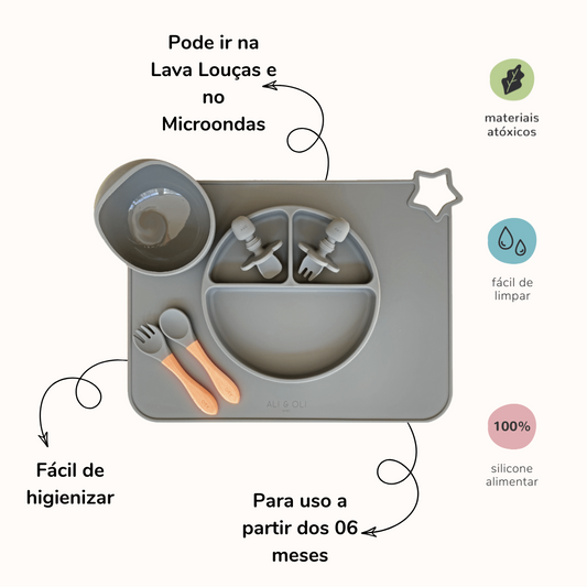 magem contem um prato de silicone com tres divisorias sendo uma maior na horizontal e duas menores na vertical. Duas colheres pequenas para bebê de silicone com limitadores. Um jogo americano retangular com uma estrela no canto esquerdo superior e o escrito Ali & Oli Baby no centro inferior. Um bowl de silicone com ventosa e duas colheres com cabo de madeira de ponteiras de silicone. Todas as peças são na cor chumbo. Imagem contem informações sobre o produto. Na esquerda: Pode ir na lava louças e no microon