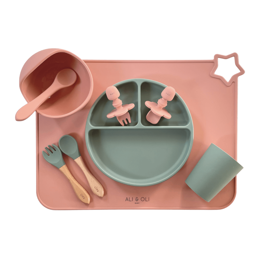 Imagem contem um Kit introdução alimentar bebê com um prato de silicone com tres divisorias sendo uma maior na horizontal e duas menores na vertical. Duas colheres pequenas para bebê de silicone com limitadores. Um jogo americano retangular com uma estrela no canto esquerdo superior e o escrito Ali & Oli Baby no centro inferior. Um bowl de silicone com ventosa e duas colheres com cabo de madeira de ponteiras de silicone, uma colher de silicone, e um copinho de silicone. As peças são umas rosa e outras verde