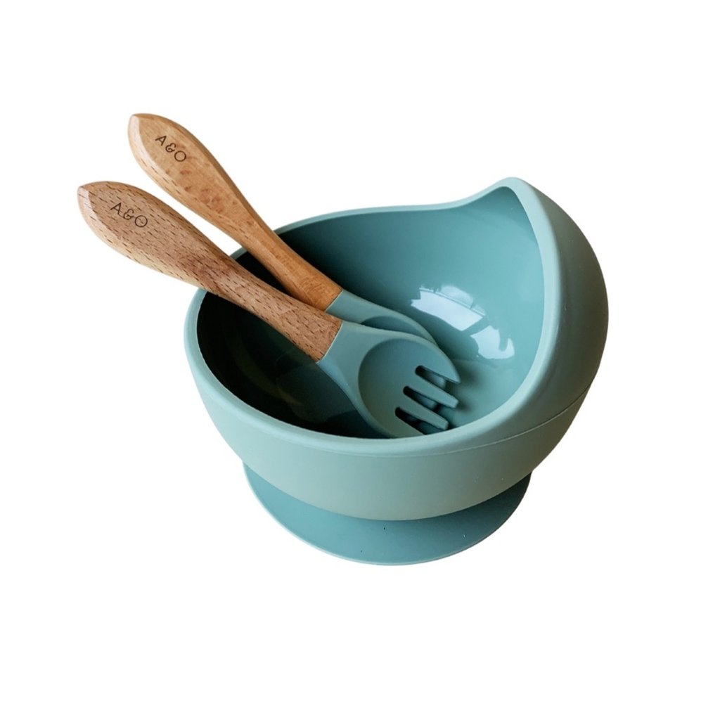 Um bowl de silicone com ventosa e duas colheres com cabo de madeira e ponteiras de silicone na cor verde sage.