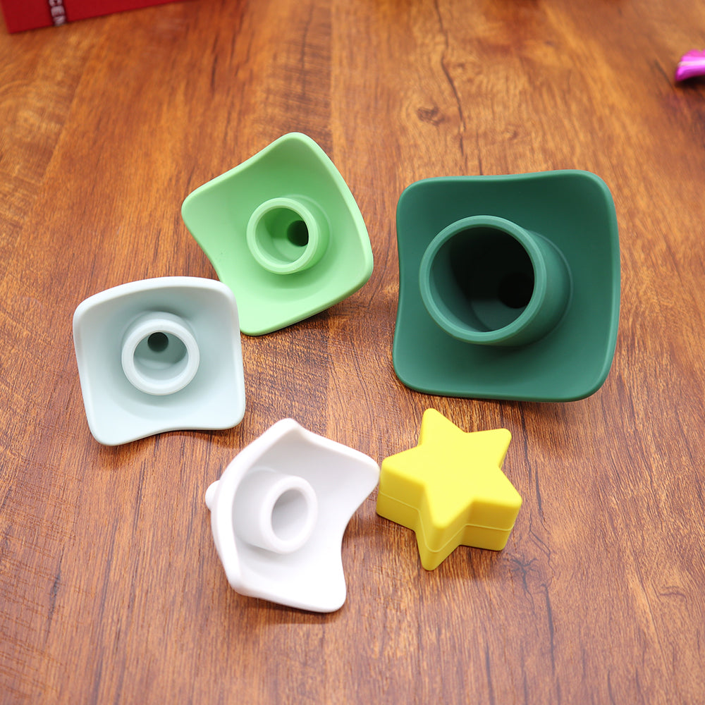 Árvore de natal em formato de peças de silicone encaixadas uma sobre a outras nas cores verde escuro, verde claro, cinza, branco e por ultimo uma estrela amarela. As peças estão desmontadas para ilustrar como é fácil a montagem da mesma para o bebê brincar.
