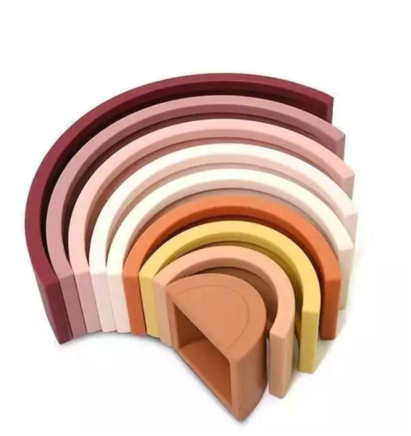 Arco-íris feito de tiras de silicone nas cores marrom, rosa, amarelo mostarda e laranja. Peças desmontáveis para brincar e empilhar e encaixar. Peças colocadas em outro modo para ilustrar como podem ser utilizados.