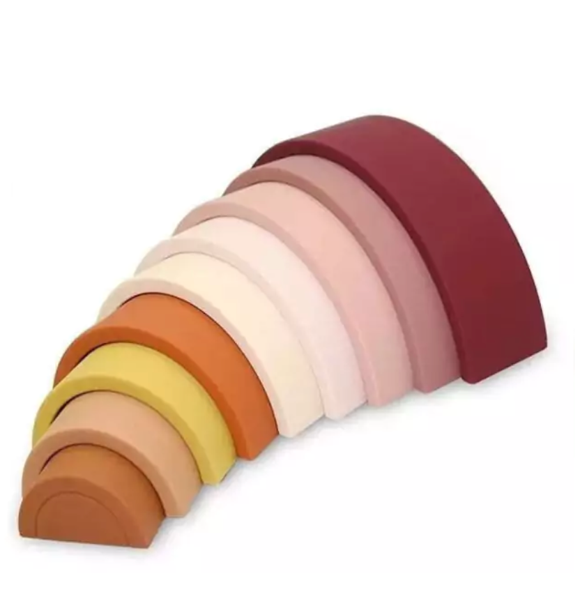 Arco-íris feito de tiras de silicone nas cores marrom, rosa, amarelo mostarda e laranja. Peças desmontáveis para brincar e empilhar e encaixar. Peças colocadas em outros modo para ilustrar como podem ser utilizadas.