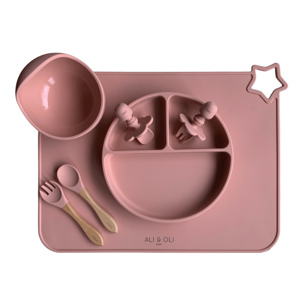 Imagem contem um Kit introdução alimentar bebê com um prato de silicone com tres divisorias sendo uma maior na horizontal e duas menores na vertical. Duas colheres pequenas para bebê de silicone com limitadores. Um jogo americano retangular com uma estrela no canto esquerdo superior e o escrito Ali & Oli Baby no centro inferior. Um bowl de silicone com ventosa e duas colheres com cabo de madeira de ponteiras de silicone. Todas as peças são na cor Rosa Velho.