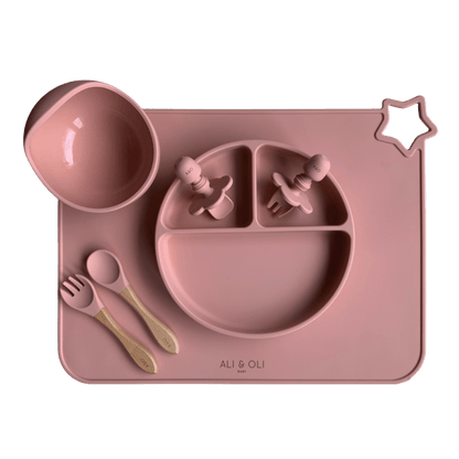 Imagem contem um Kit introdução alimentar bebê com um prato de silicone com tres divisorias sendo uma maior na horizontal e duas menores na vertical. Duas colheres pequenas para bebê de silicone com limitadores. Um jogo americano retangular com uma estrela no canto esquerdo superior e o escrito Ali & Oli Baby no centro inferior. Um bowl de silicone com ventosa e duas colheres com cabo de madeira de ponteiras de silicone. Todas as peças são na cor Rosa Velho.