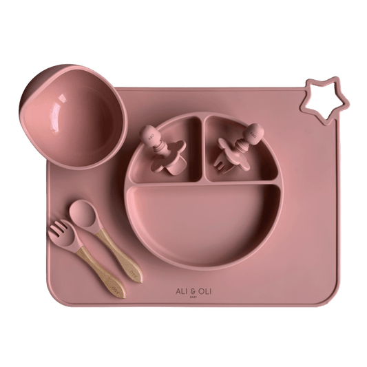 Imagem contem um Kit introdução alimentar bebê com um prato de silicone com tres divisorias sendo uma maior na horizontal e duas menores na vertical. Duas colheres pequenas para bebê de silicone com limitadores. Um jogo americano retangular com uma estrela no canto esquerdo superior e o escrito Ali & Oli Baby no centro inferior. Um bowl de silicone com ventosa e duas colheres com cabo de madeira de ponteiras de silicone. Todas as peças são na cor Rosa Velho.