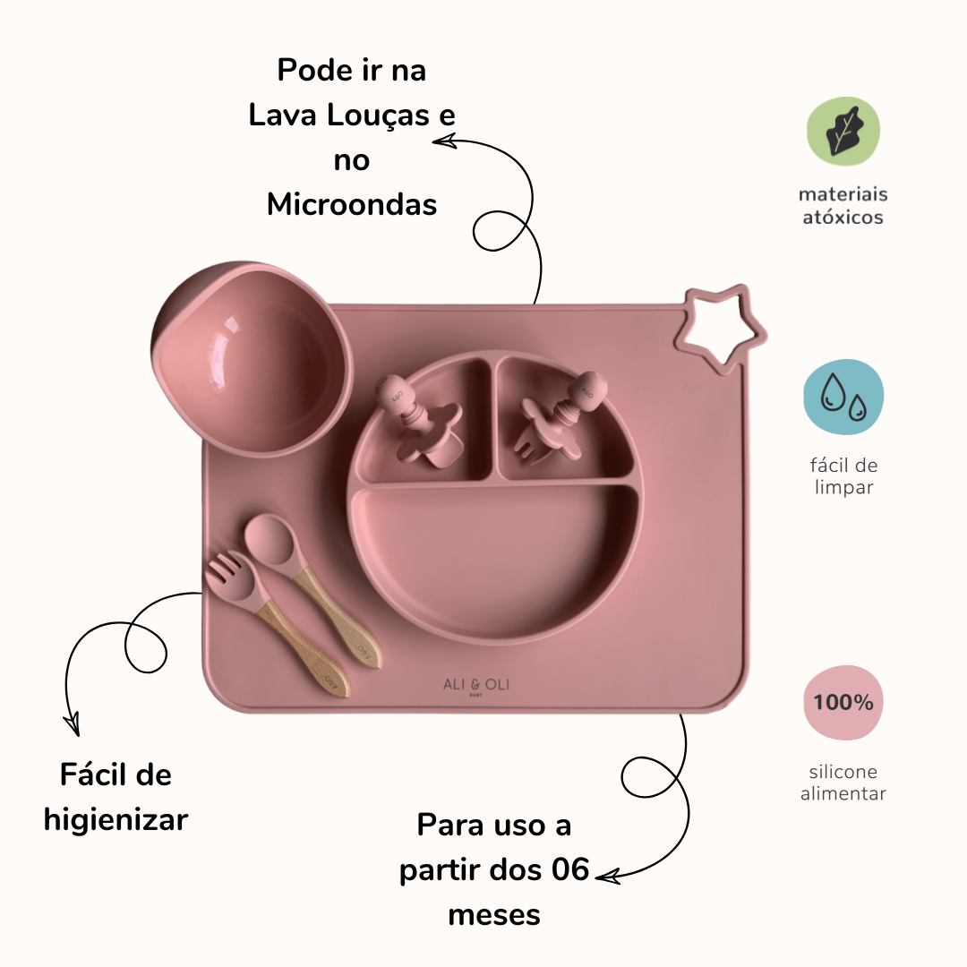 Imagem contem um prato de silicone com tres divisorias sendo uma maior na horizontal e duas menores na vertical. Duas colheres pequenas para bebê de silicone com limitadores. Um jogo americano retangular com uma estrela no canto esquerdo superior e o escrito Ali & Oli Baby no centro inferior. Um bowl de silicone com ventosa e duas colheres com cabo de madeira de ponteiras de silicone. Todas as peças são na cor Rosa Velho. Imagem contem informações sobre o produto. Na esquerda: Pode ir na lava louças e no mi