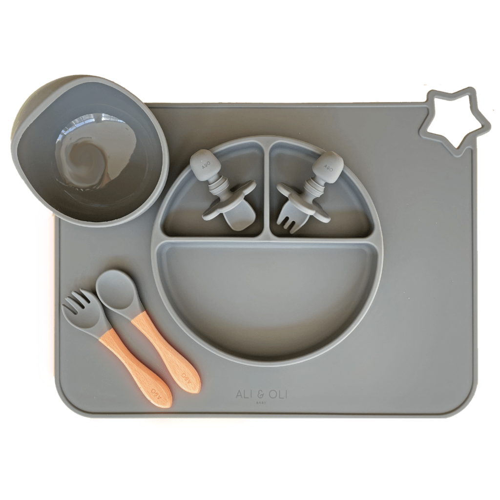 Imagem contem um Kit introdução alimentar bebê com um prato de silicone com tres divisorias sendo uma maior na horizontal e duas menores na vertical. Duas colheres pequenas para bebê de silicone com limitadores. Um jogo americano retangular com uma estrela no canto esquerdo superior e o escrito Ali & Oli Baby no centro inferior. Um bowl de silicone com ventosa e duas colheres com cabo de madeira de ponteiras de silicone. Todas as peças são na cor chumbo.