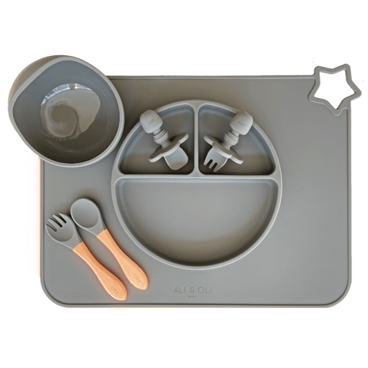 Imagem contem um Kit introdução alimentar bebê com um prato de silicone com tres divisorias sendo uma maior na horizontal e duas menores na vertical. Duas colheres pequenas para bebê de silicone com limitadores. Um jogo americano retangular com uma estrela no canto esquerdo superior e o escrito Ali & Oli Baby no centro inferior. Um bowl de silicone com ventosa e duas colheres com cabo de madeira de ponteiras de silicone. Todas as peças são na cor chumbo.