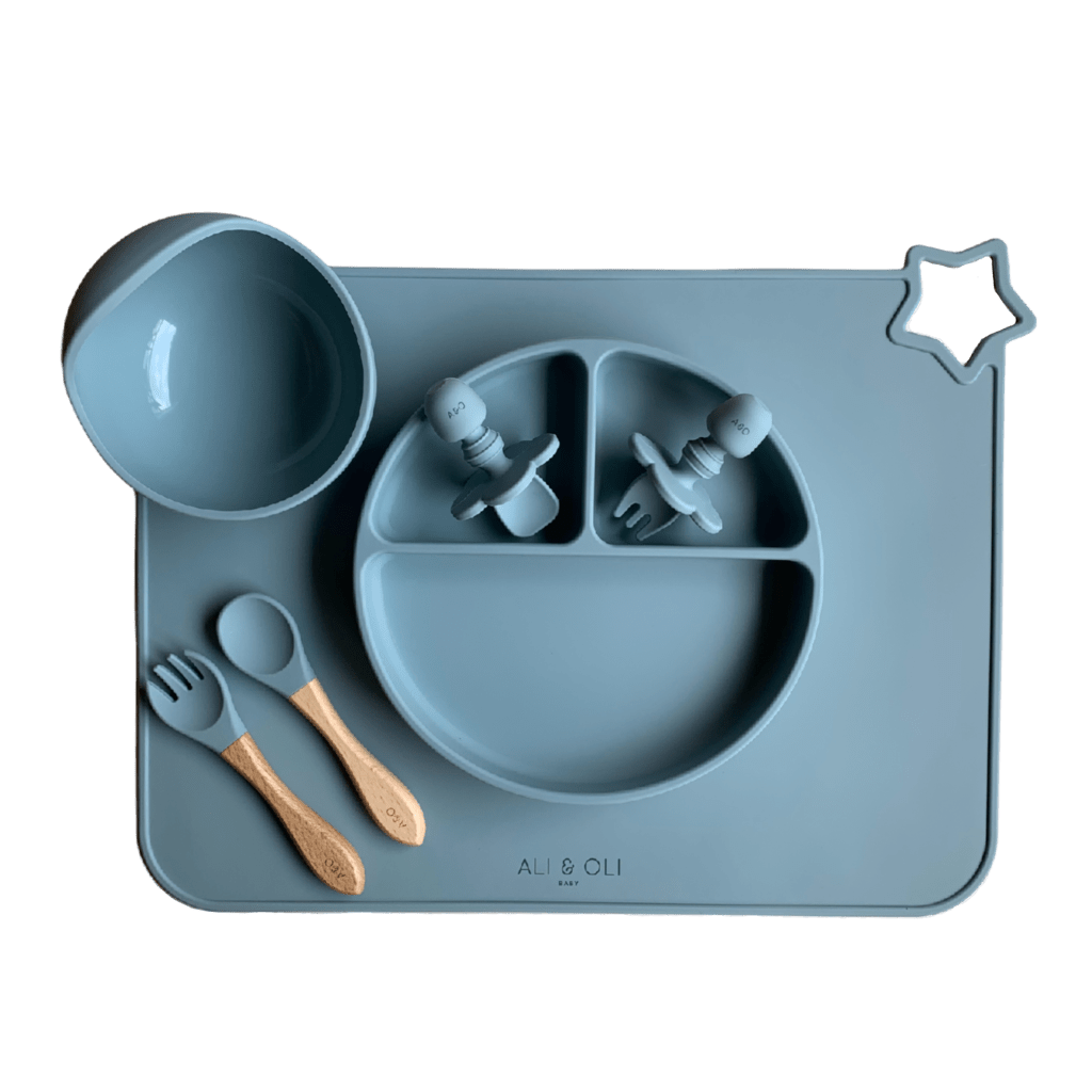 Imagem contem um Kit introdução alimentar bebê com um prato de silicone com tres divisorias sendo uma maior na horizontal e duas menores na vertical. Duas colheres pequenas para bebê de silicone com limitadores. Um jogo americano retangular com uma estrela no canto esquerdo superior e o escrito Ali & Oli Baby no centro inferior. Um bowl de silicone com ventosa e duas colheres com cabo de madeira de ponteiras de silicone. Todas as peças são na cor Azul Ether