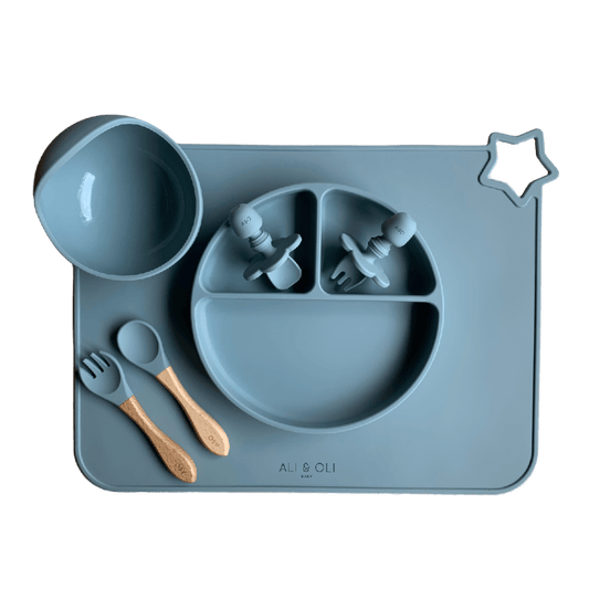 Imagem contem um Kit introdução alimentar bebê com um prato de silicone com tres divisorias sendo uma maior na horizontal e duas menores na vertical. Duas colheres pequenas para bebê de silicone com limitadores. Um jogo americano retangular com uma estrela no canto esquerdo superior e o escrito Ali & Oli Baby no centro inferior. Um bowl de silicone com ventosa e duas colheres com cabo de madeira de ponteiras de silicone. Todas as peças são na cor Azul Ether