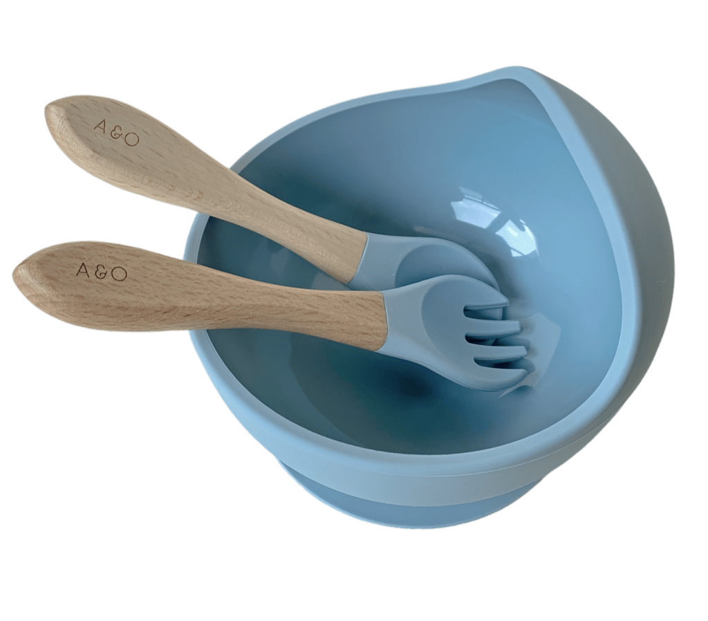 Um bowl de silicone com ventosa e duas colheres com cabo de madeira e ponteiras de silicone na cor azul ether.