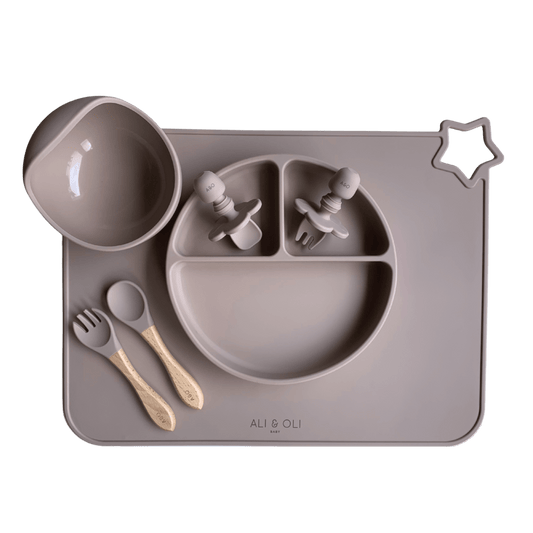Imagem contem um Kit introdução alimentar bebê com um prato de silicone com tres divisorias sendo uma maior na horizontal e duas menores na vertical. Duas colheres pequenas para bebê de silicone com limitadores. Um jogo americano retangular com uma estrela no canto esquerdo superior e o escrito Ali & Oli Baby no centro inferior. Um bowl de silicone com ventosa e duas colheres com cabo de madeira de ponteiras de silicone. Todas as peças são na cor nude.