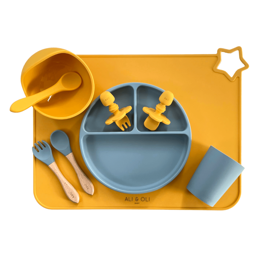Imagem contem um Kit introdução alimentar bebê com um prato de silicone com tres divisorias sendo uma maior na horizontal e duas menores na vertical. Duas colheres pequenas para bebê de silicone com limitadores. Um jogo americano retangular com uma estrela no canto esquerdo superior e o escrito Ali & Oli Baby no centro inferior. Um bowl de silicone com ventosa e duas colheres com cabo de madeira de ponteiras de silicone, uma colher de silicone, e um copinho de silicone. As peças são amarelas ou azuis.