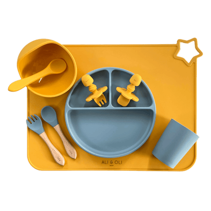Imagem contem um Kit introdução alimentar bebê com um prato de silicone com tres divisorias sendo uma maior na horizontal e duas menores na vertical. Duas colheres pequenas para bebê de silicone com limitadores. Um jogo americano retangular com uma estrela no canto esquerdo superior e o escrito Ali & Oli Baby no centro inferior. Um bowl de silicone com ventosa e duas colheres com cabo de madeira de ponteiras de silicone, uma colher de silicone, e um copinho de silicone. As peças são amarelas ou azuis.