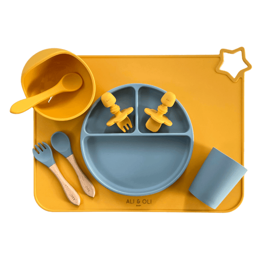 Imagem contem um Kit introdução alimentar bebê com um prato de silicone com tres divisorias sendo uma maior na horizontal e duas menores na vertical. Duas colheres pequenas para bebê de silicone com limitadores. Um jogo americano retangular com uma estrela no canto esquerdo superior e o escrito Ali & Oli Baby no centro inferior. Um bowl de silicone com ventosa e duas colheres com cabo de madeira de ponteiras de silicone, uma colher de silicone, e um copinho de silicone. As peças são amarelas ou azuis.