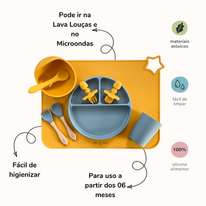 Imagem contem um Kit introdução alimentar bebê com um prato de silicone com tres divisorias sendo uma maior na horizontal e duas menores na vertical. Duas colheres pequenas para bebê de silicone com limitadores. Um jogo americano retangular com uma estrela no canto esquerdo superior e o escrito Ali & Oli Baby no centro inferior. Um bowl de silicone com ventosa e duas colheres com cabo de madeira de ponteiras de silicone, uma colher de silicone, e um copinho de silicone. As peças são amarelas ou azuis.