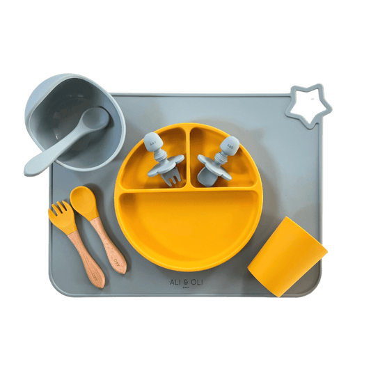 Imagem contem um Kit introdução alimentar bebê com um prato de silicone com tres divisorias sendo uma maior na horizontal e duas menores na vertical. Duas colheres pequenas para bebê de silicone com limitadores. Um jogo americano retangular com uma estrela no canto esquerdo superior e o escrito Ali & Oli Baby no centro inferior. Um bowl de silicone com ventosa e duas colheres com cabo de madeira de ponteiras de silicone, uma colher de silicone, e um copinho de silicone. As peças são umas azul e outras amare