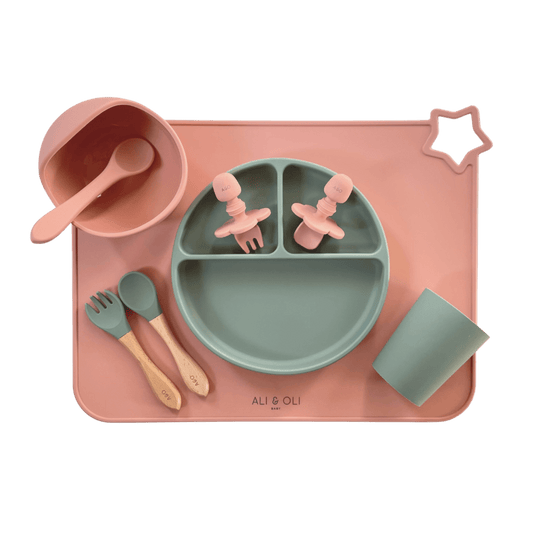 Imagem contem um Kit introdução alimentar bebê com um prato de silicone com tres divisorias sendo uma maior na horizontal e duas menores na vertical. Duas colheres pequenas para bebê de silicone com limitadores. Um jogo americano retangular com uma estrela no canto esquerdo superior e o escrito Ali & Oli Baby no centro inferior. Um bowl de silicone com ventosa e duas colheres com cabo de madeira de ponteiras de silicone, uma colher de silicone, e um copinho de silicone. As peças são umas rosa e outras verde