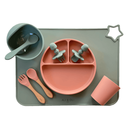 Imagem contem um Kit introdução alimentar bebê com um prato de silicone com tres divisorias sendo uma maior na horizontal e duas menores na vertical. Duas colheres pequenas para bebê de silicone com limitadores. Um jogo americano retangular com uma estrela no canto esquerdo superior e o escrito Ali & Oli Baby no centro inferior. Um bowl de silicone com ventosa e duas colheres com cabo de madeira de ponteiras de silicone, uma colher de silicone, e um copinho de silicone. As peças são umas rosa e outras verde