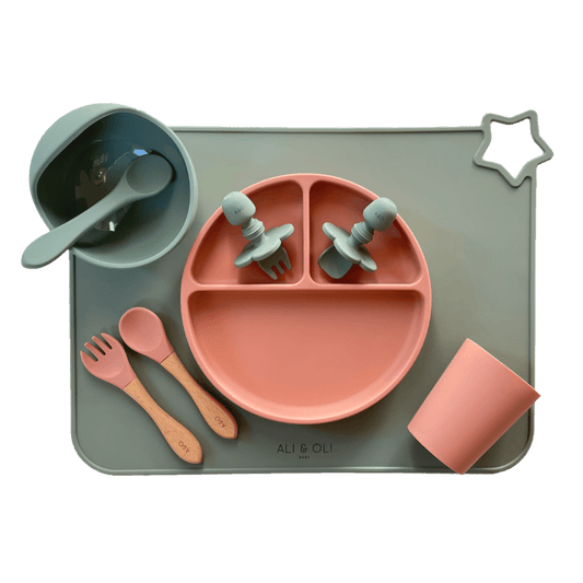Imagem contem um Kit introdução alimentar bebê com um prato de silicone com tres divisorias sendo uma maior na horizontal e duas menores na vertical. Duas colheres pequenas para bebê de silicone com limitadores. Um jogo americano retangular com uma estrela no canto esquerdo superior e o escrito Ali & Oli Baby no centro inferior. Um bowl de silicone com ventosa e duas colheres com cabo de madeira de ponteiras de silicone, uma colher de silicone, e um copinho de silicone. As peças são umas rosa e outras verde