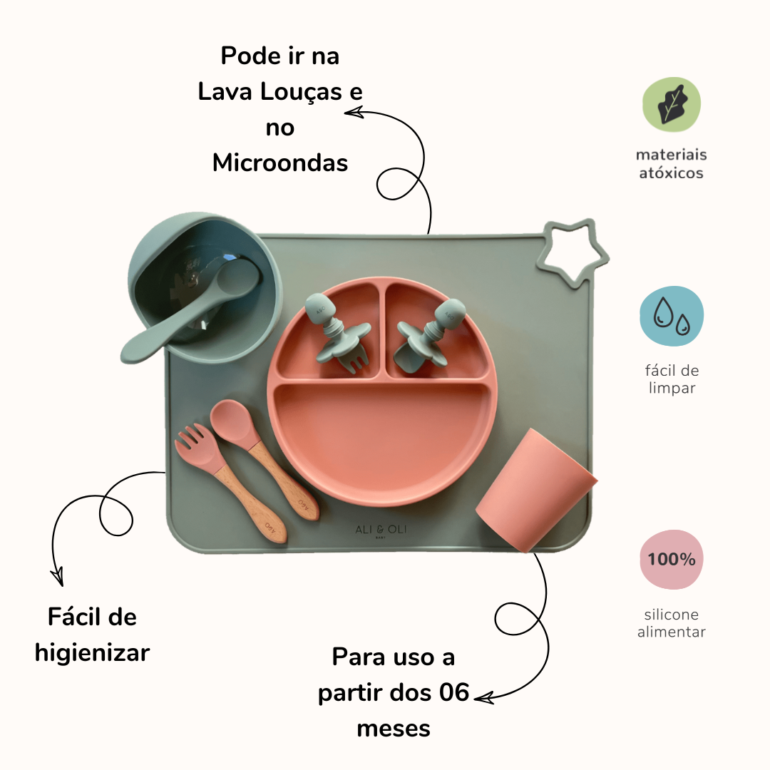 Imagem contem um Kit introdução alimentar bebê com um prato de silicone com tres divisorias sendo uma maior na horizontal e duas menores na vertical. Duas colheres pequenas para bebê de silicone com limitadores. Um jogo americano retangular com uma estrela no canto esquerdo superior e o escrito Ali & Oli Baby no centro inferior. Um bowl de silicone com ventosa e duas colheres com cabo de madeira de ponteiras de silicone, uma colher de silicone, e um copinho de silicone. As peças são umas rosa e outras verde