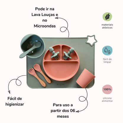 Imagem contem um Kit introdução alimentar bebê com um prato de silicone com tres divisorias sendo uma maior na horizontal e duas menores na vertical. Duas colheres pequenas para bebê de silicone com limitadores. Um jogo americano retangular com uma estrela no canto esquerdo superior e o escrito Ali & Oli Baby no centro inferior. Um bowl de silicone com ventosa e duas colheres com cabo de madeira de ponteiras de silicone, uma colher de silicone, e um copinho de silicone. As peças são umas rosa e outras verde