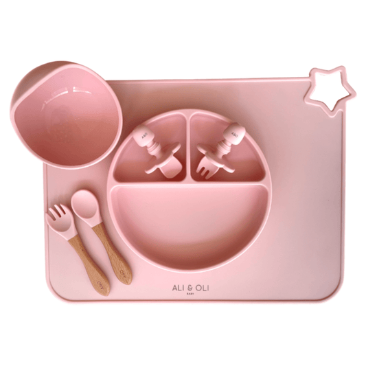 Imagem contem um Kit introdução alimentar bebê com um prato de silicone com tres divisorias sendo uma maior na horizontal e duas menores na vertical. Duas colheres pequenas para bebê de silicone com limitadores. Um jogo americano retangular com uma estrela no canto esquerdo superior e o escrito Ali & Oli Baby no centro inferior. Um bowl de silicone com ventosa e duas colheres com cabo de madeira de ponteiras de silicone. Todas as peças são na cor Rosa bebê. 