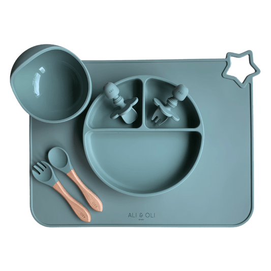 silicone com tres divisorias sendo uma maior na horizontal e duas menores na vertical. Duas colheres pequenas para bebê de silicone com limitadores. Um jogo americano retangular com uma estrela no canto esquerdo superior e o escrito Ali & Oli Baby no centro inferior. Um bowl de silicone com ventosa e duas colheres com cabo de madeira de ponteiras de silicone. Todas as peças são na cor Verde Sage.