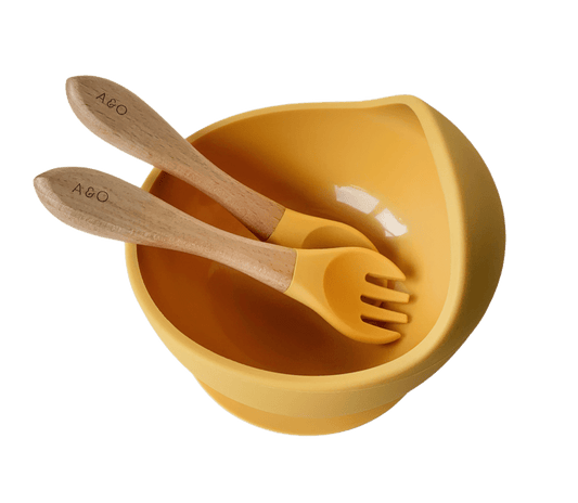 Um bowl com uma colher e um garfo com cabo de madeira e ponteira de silicone na cor amarelo mostarda.